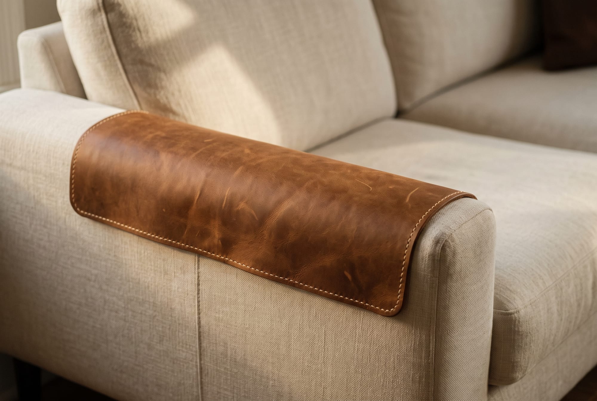 A tobacco crazy horse leather armrest panel running the full length of a cream linen sofa arm, its edge falling at the seam line rather than in the middle of the panel