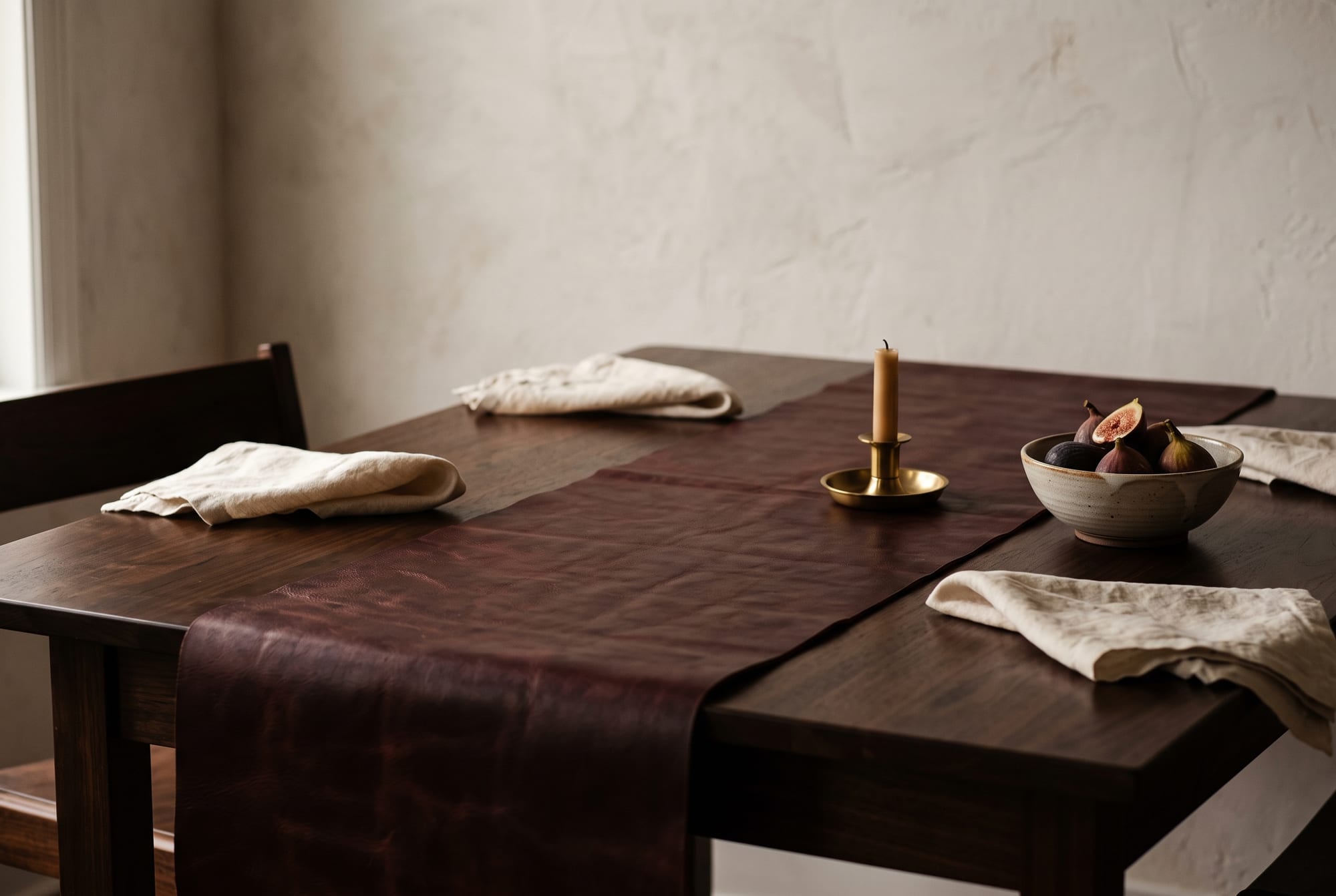 An oxblood full-grain leather table runner laid along a dark walnut dining table with cream linen napkins, a brass candle holder and autumn window light