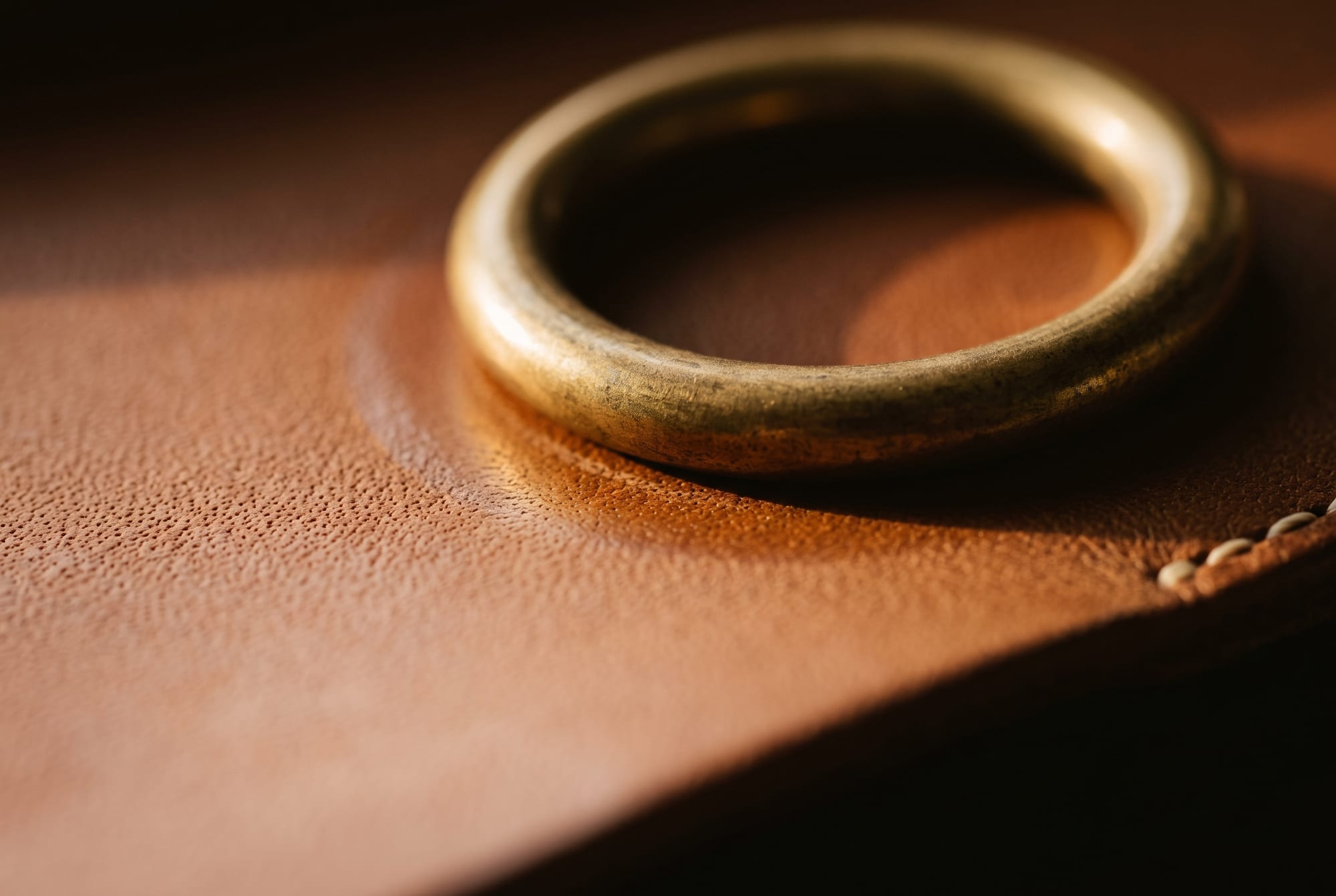 Macro of an unlacquered brass spring ring resting against cognac full-grain leather, the metal showing a warm darkened patina and the leather beneath it faintly polished where the two have been in contact