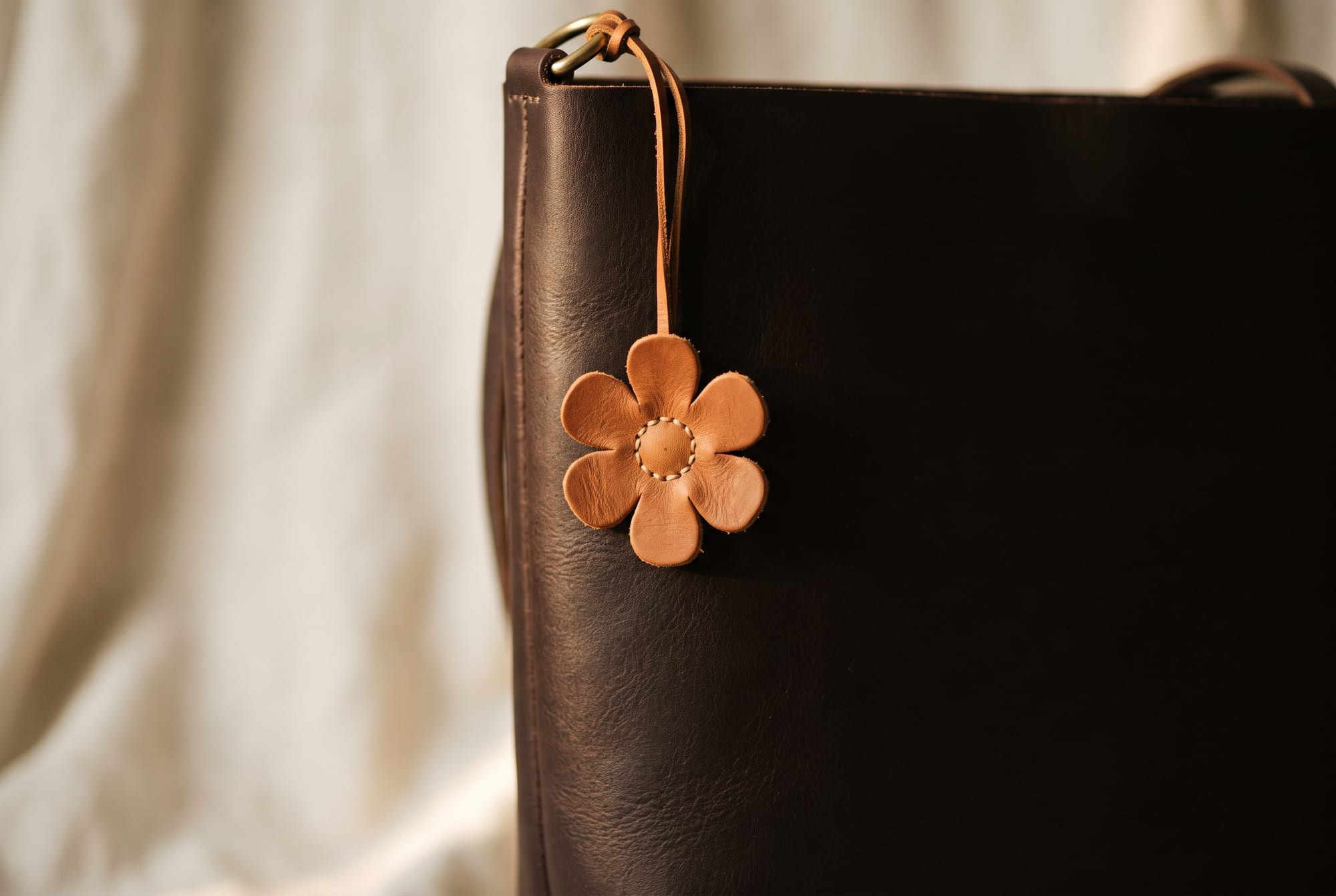 A single hand-stitched cognac leather flower charm hanging from the side ring of an otherwise plain chocolate leather tote against a cream linen background, with generous empty space around it