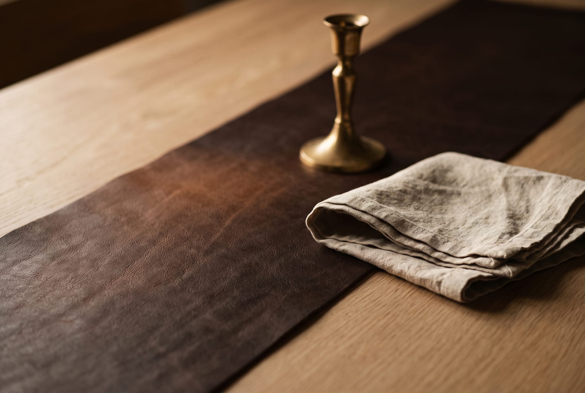 A chocolate brown leather table runner on a pale oak dining table beside an oatmeal linen napkin and an unlacquered brass candlestick, warm low window light raking across the grain