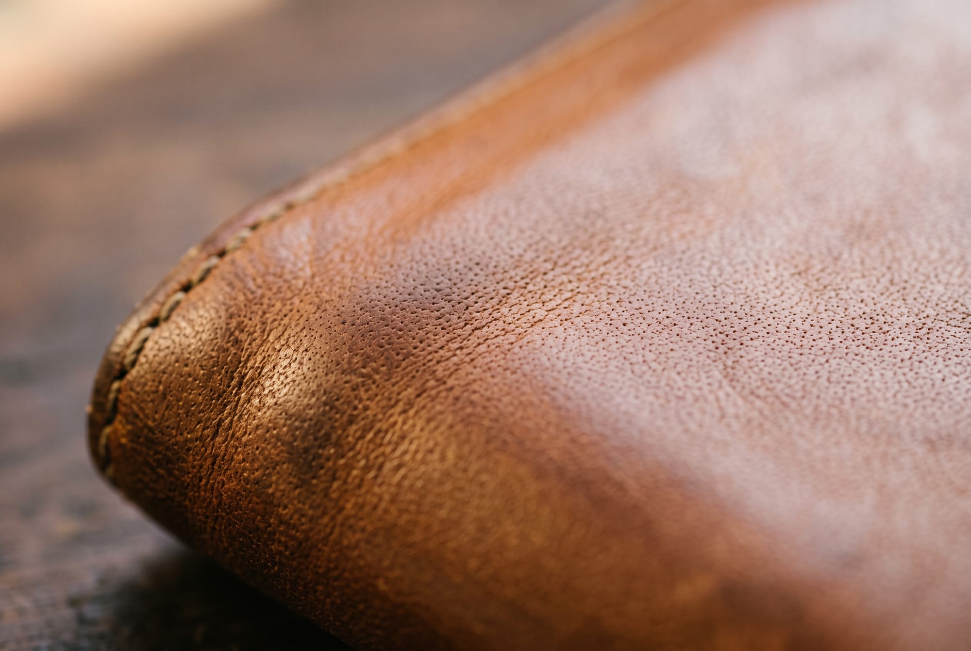 Extreme macro of the thumb corner of a cognac leather wallet, the leather distinctly darker and slightly glossy in a soft-edged halo where a thumb has rested for years, open grain pores still visible at the edge of the darkening