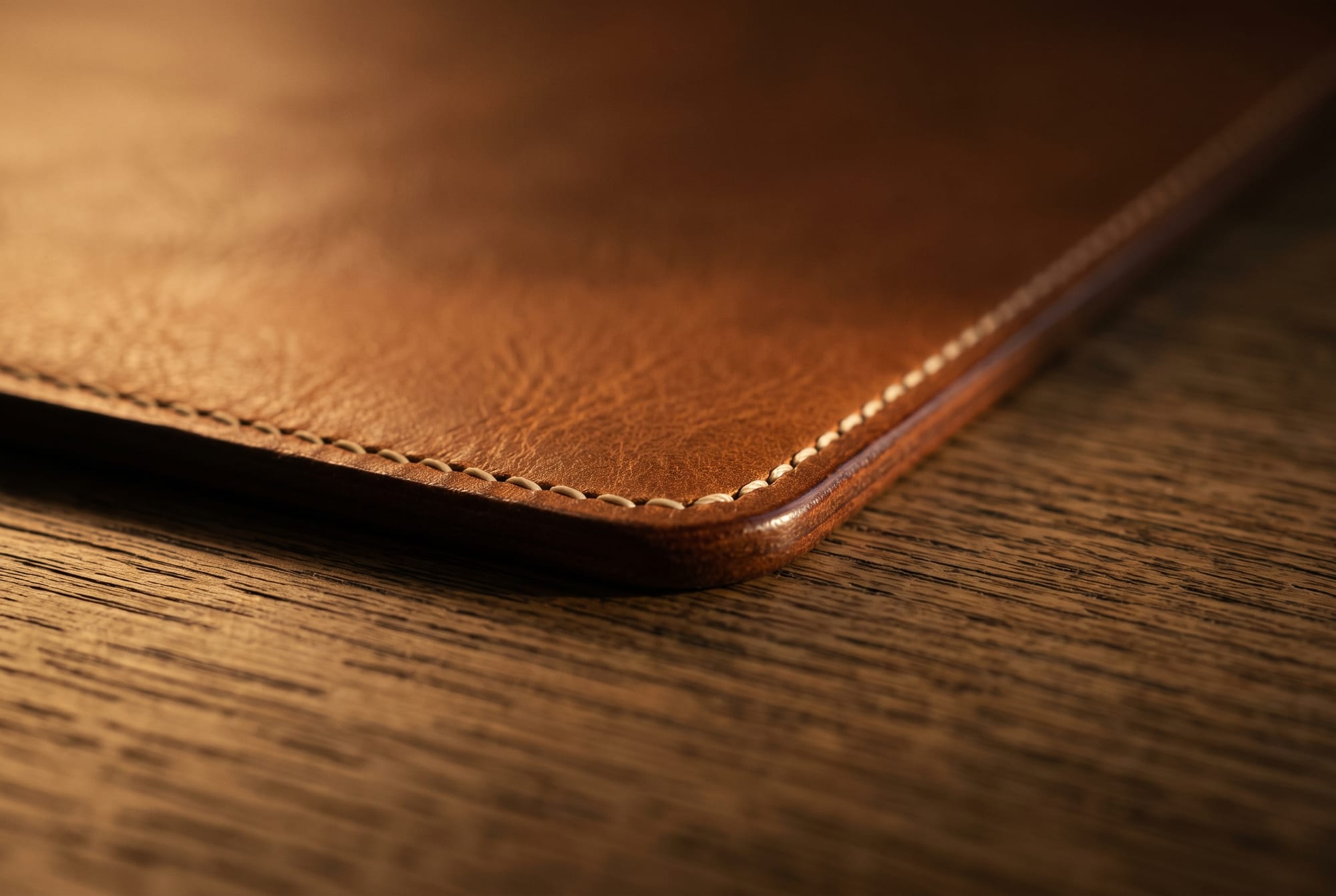 Close detail of the corner of a thick vegetable-tanned leather desk pad lying perfectly flat on oak, its burnished cut edge and hand-stitched corner visible, with the grain of the wood showing in the margin beyond