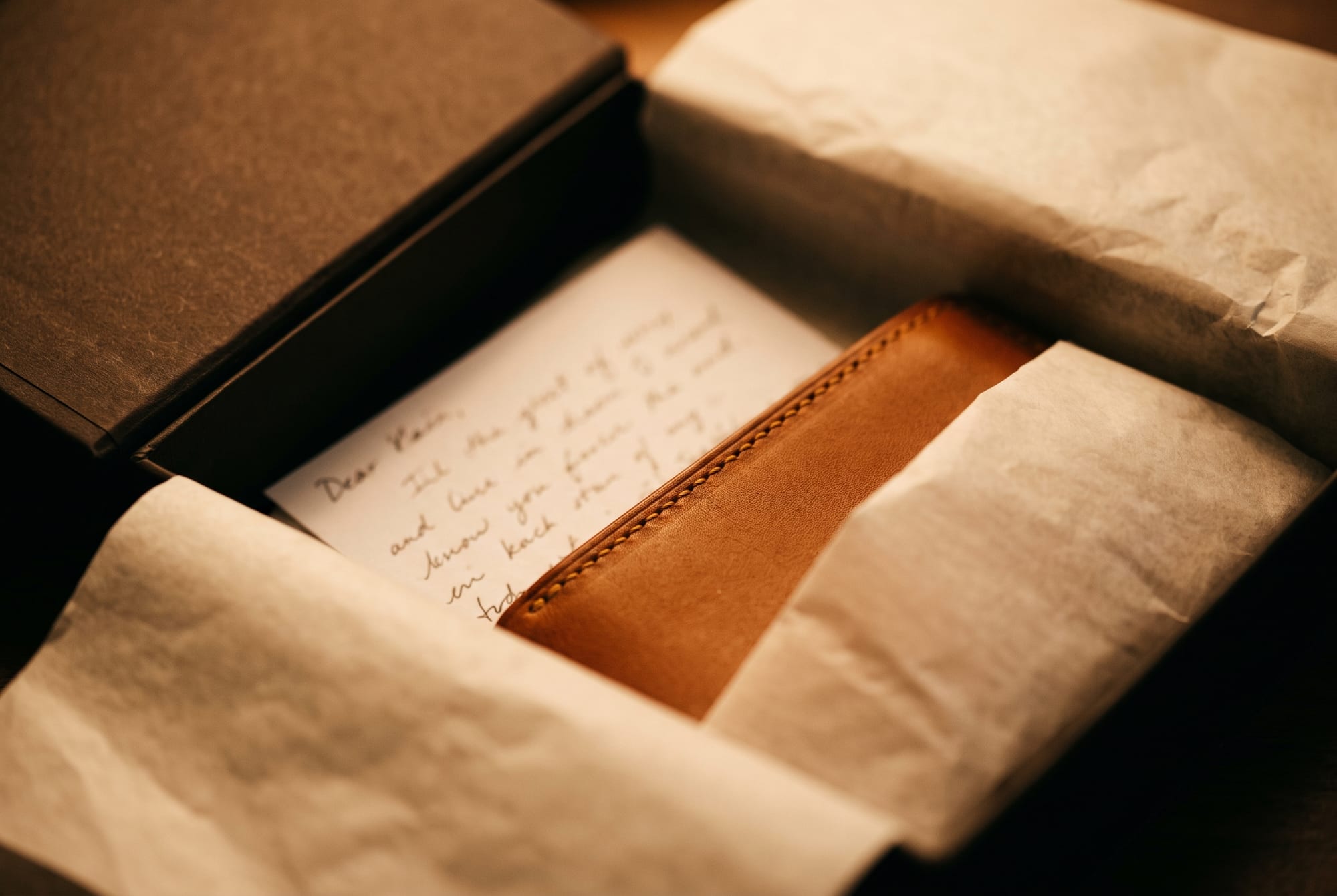 A small handwritten card resting on folded tissue inside an open gift box beside a hand-stitched leather piece, the handwriting soft-focused
