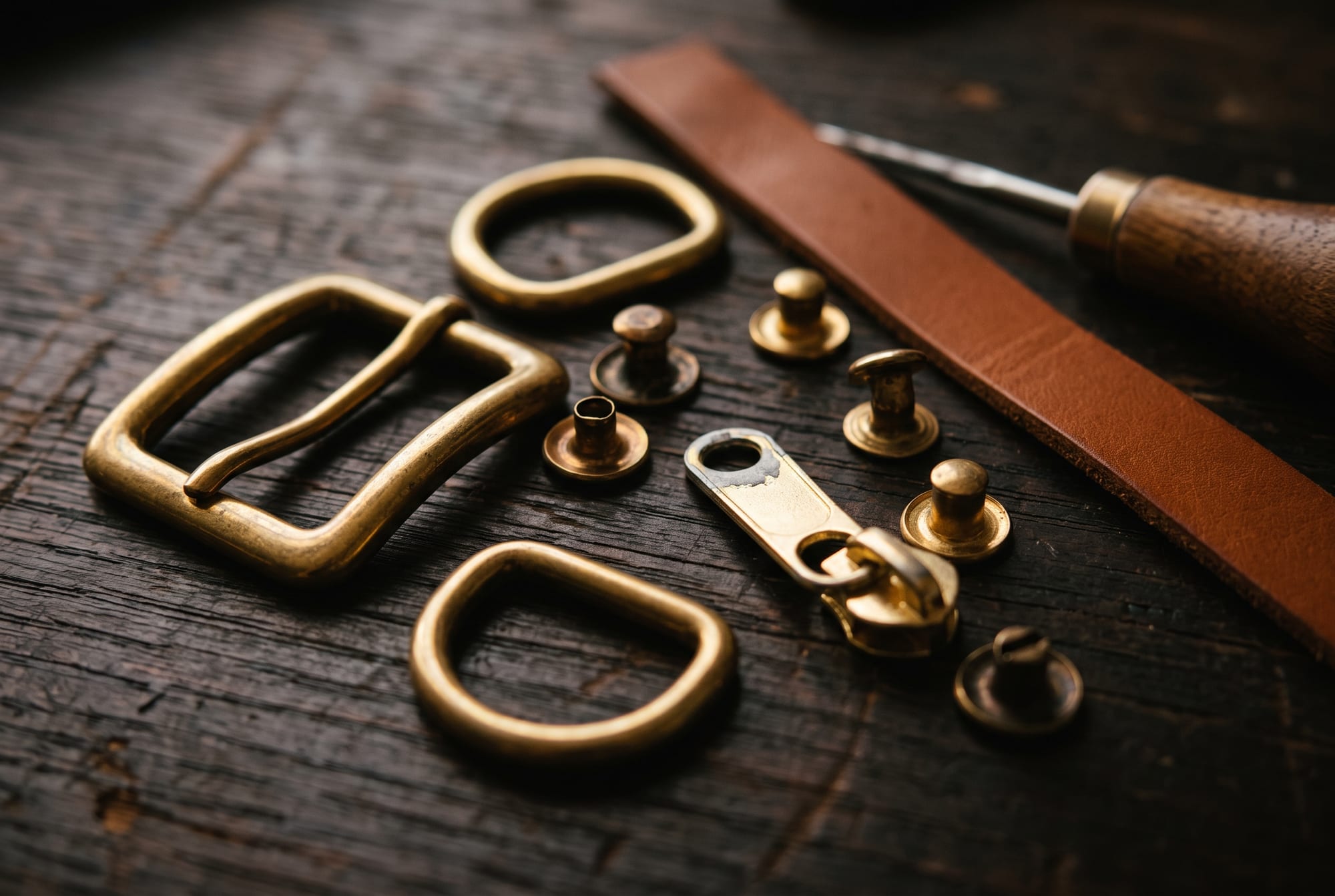 An assortment of bag fittings laid out on a worn workbench in raking light: solid brass rivets and buckles with a warm tarnished glow beside a bright plated slider showing grey metal at its worn edge, with a strip of cognac leather and a stitching awl alongside