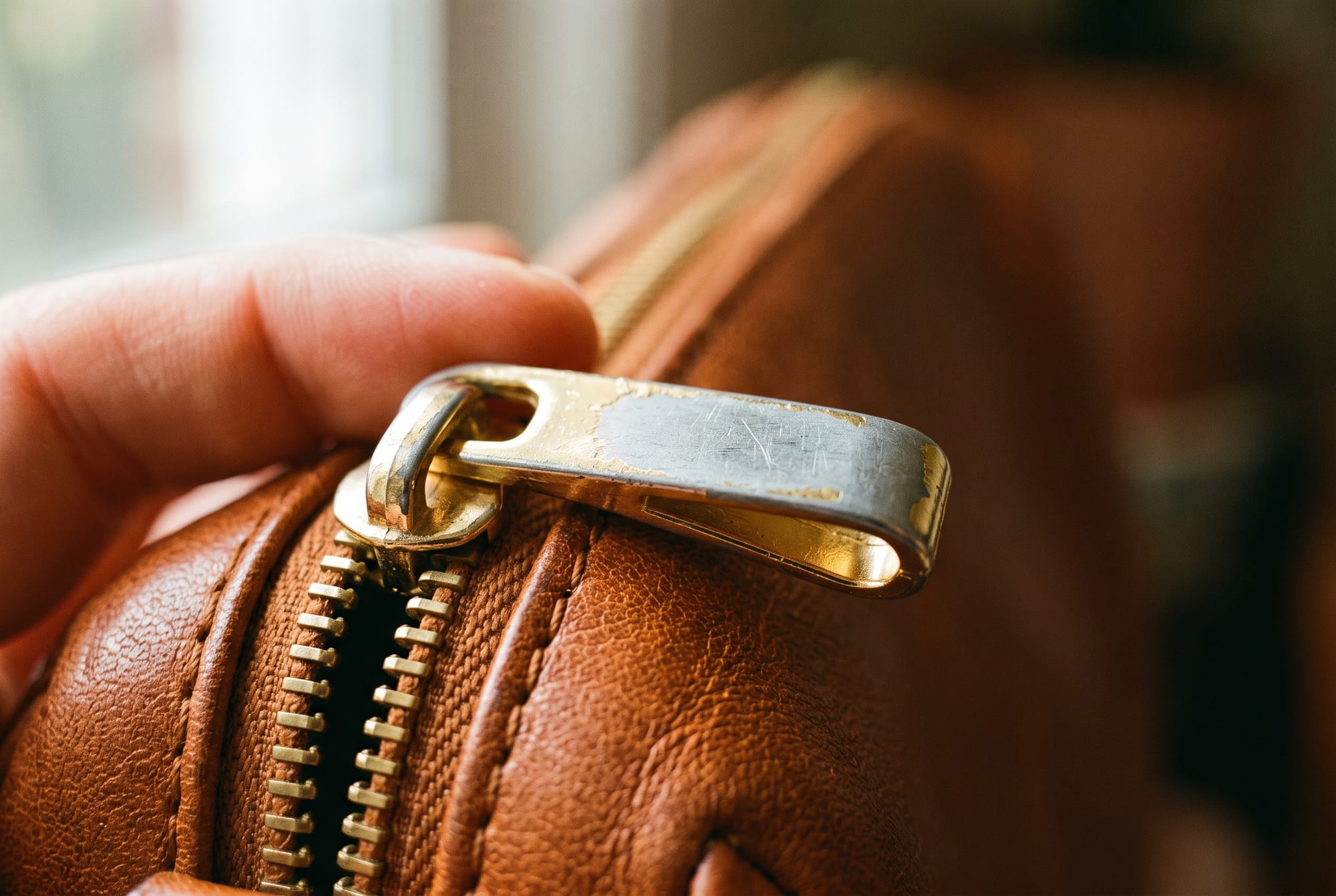 Macro photograph of a zipper pull on a cognac leather bag where the bright plating has worn through at the thumb contact point, exposing dull grey metal beneath, with the leather around it in good condition