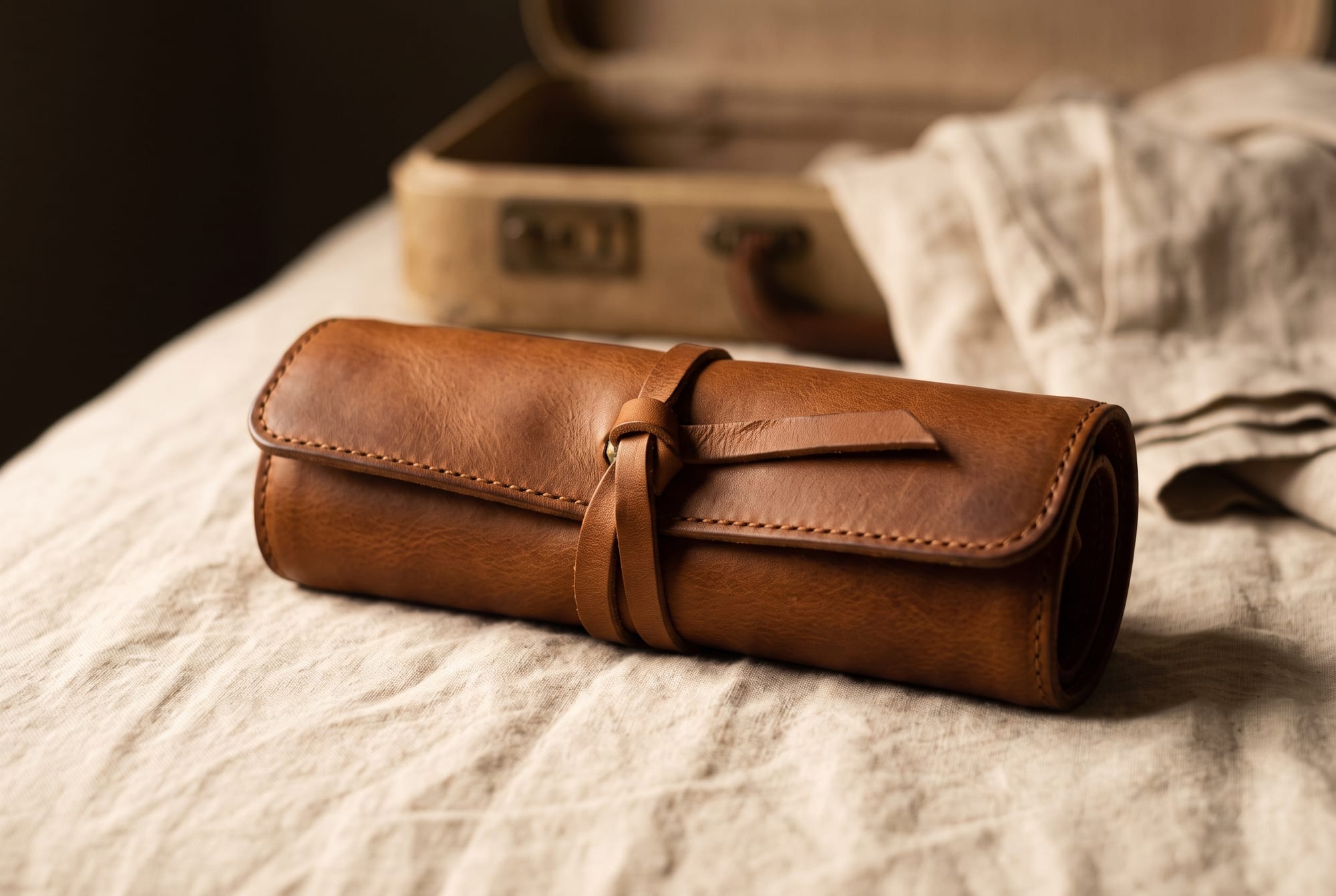 A cognac leather jewellery roll tied closed with a leather strap, resting on a linen-covered surface beside an open suitcase corner in warm low light