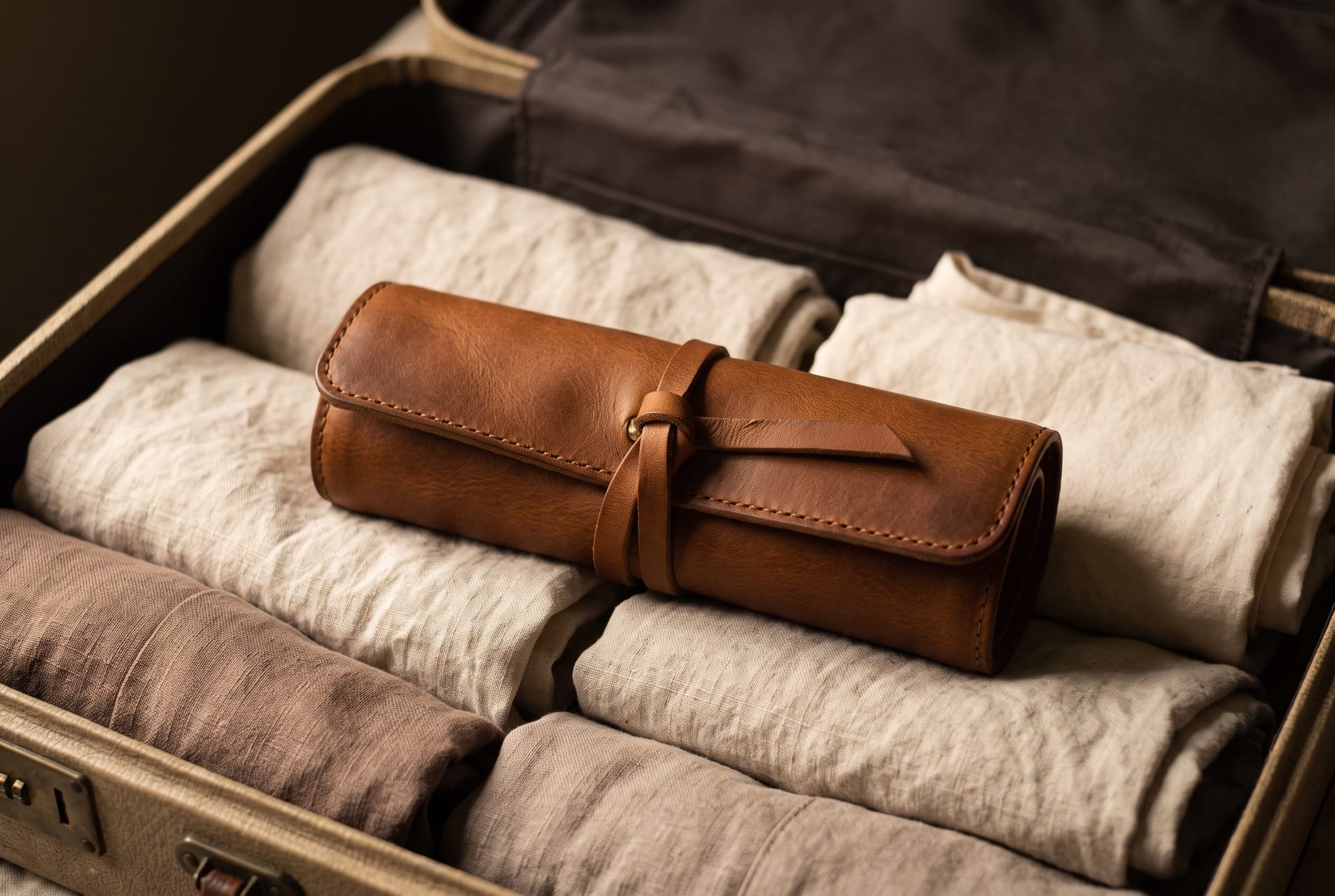 A cognac leather jewellery roll rolled closed and tied, laid flat in an open carry-on suitcase between folded linen garments, the outer leather visibly darker and softly scuffed from use