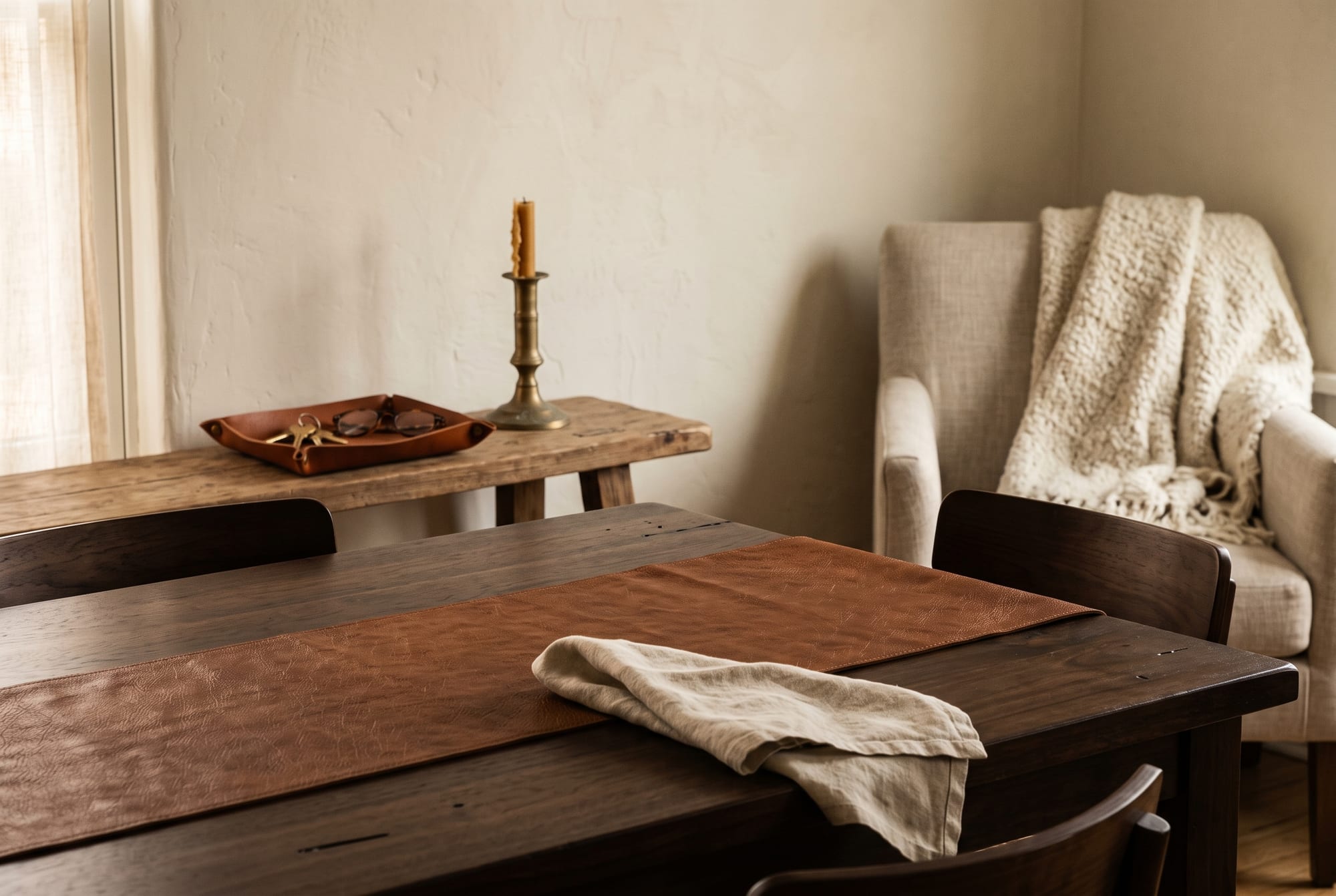 An autumn living-room corner in late afternoon light: a walnut table with a cognac leather runner, an oatmeal linen sofa, a marble surface holding a woven leather catchall, aged brass and matte ceramic