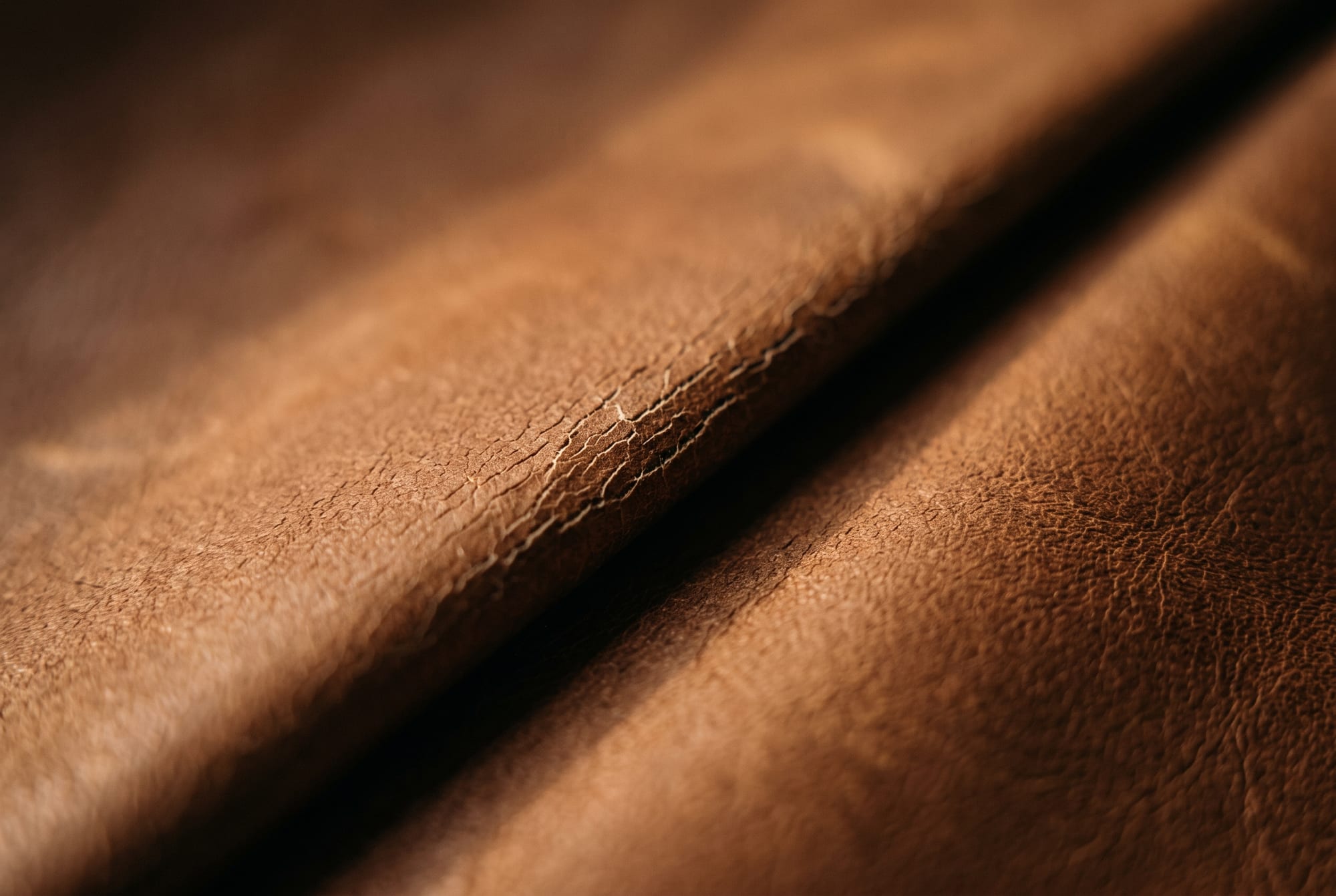 An extreme close-up of a leather fold under raking light, showing fine pale cracks opening along the crease line where the grain has broken