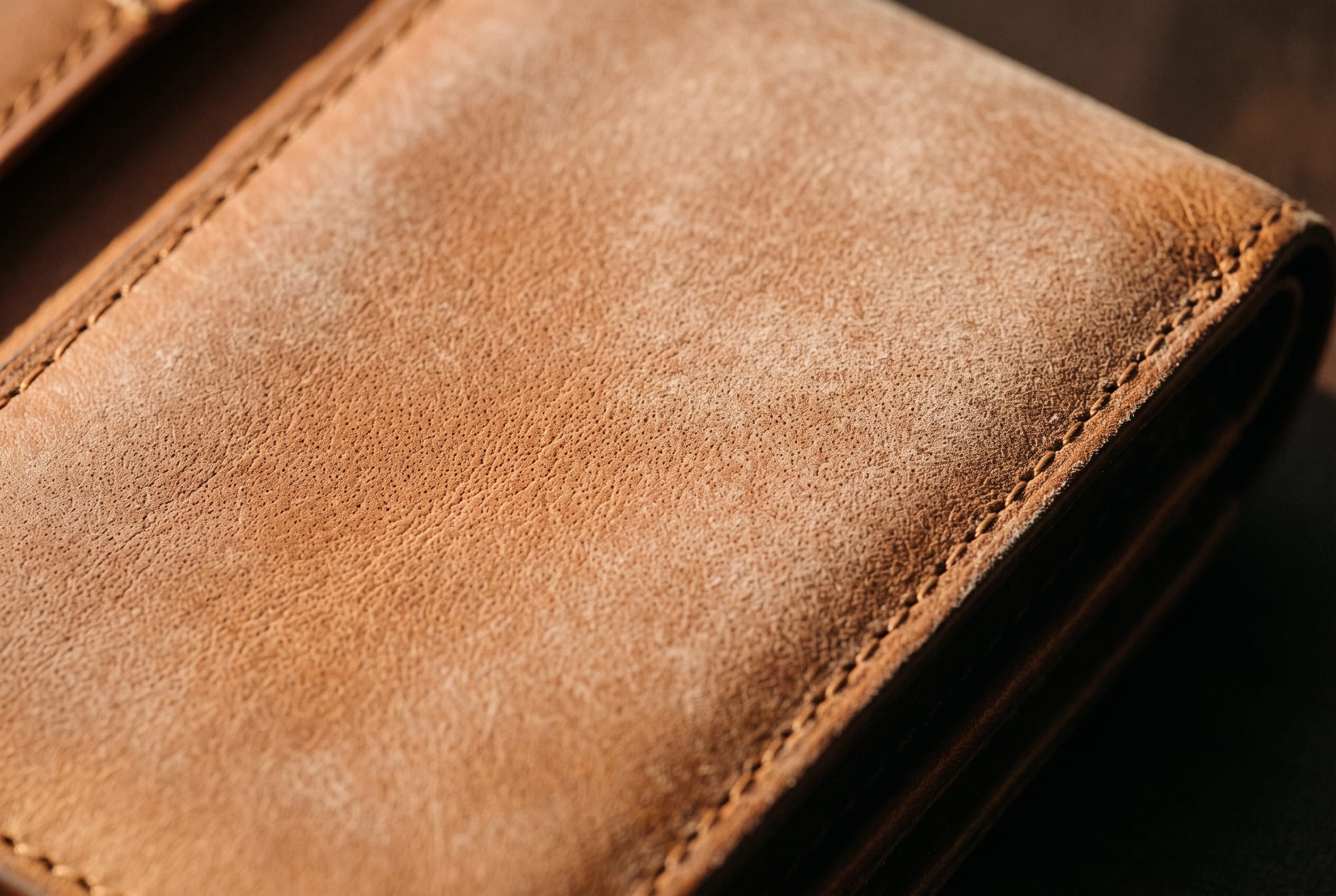 Raking low light across a cognac leather flap showing an even, dry, waxy white haze lying flat on the surface with no fuzz at its edge, the grain pores still clearly visible through it