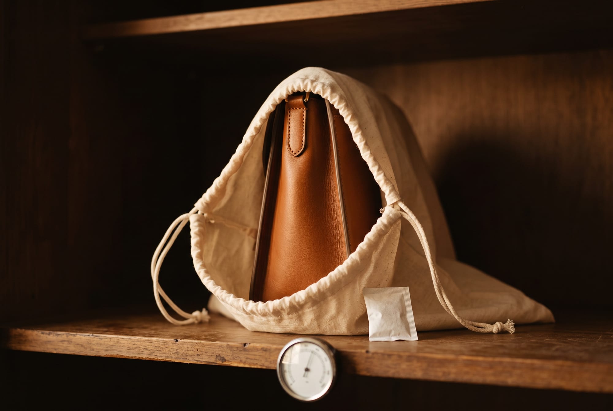 A cognac leather bag standing upright on a wooden shelf in a breathable cream cotton dust bag, clear air gaps on both sides and above it, with a small silica gel sachet tucked beside it and a dial hygrometer on the shelf edge