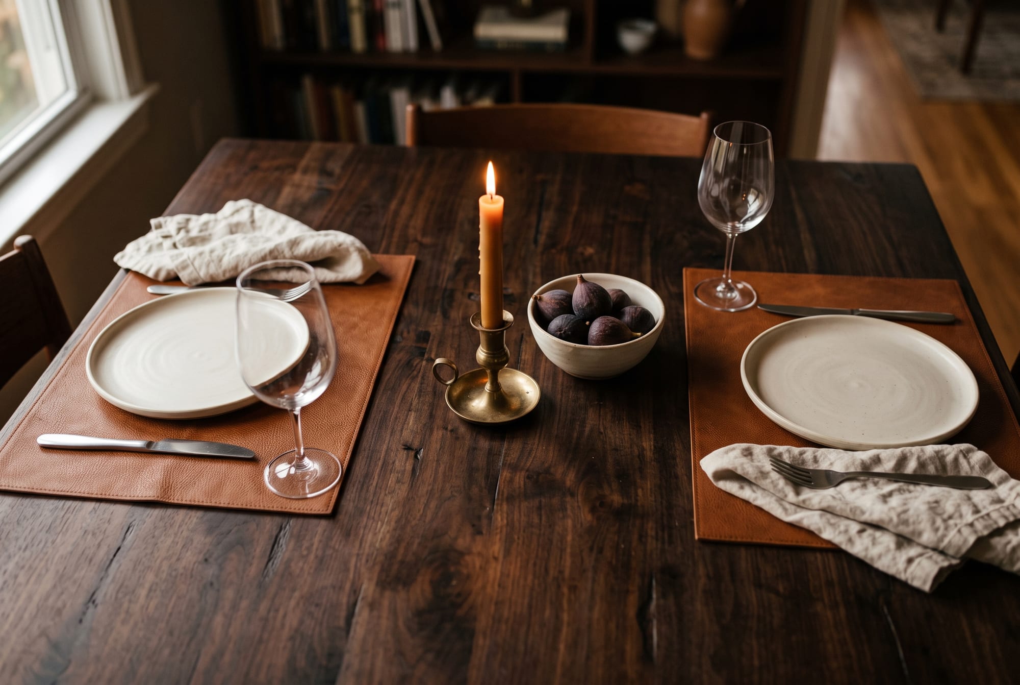 An autumn dinner table photographed from above in warm evening light: two tobacco-brown leather placemats on dark walnut, cream linen napkins, matte ceramic plates and an unlacquered brass candlestick