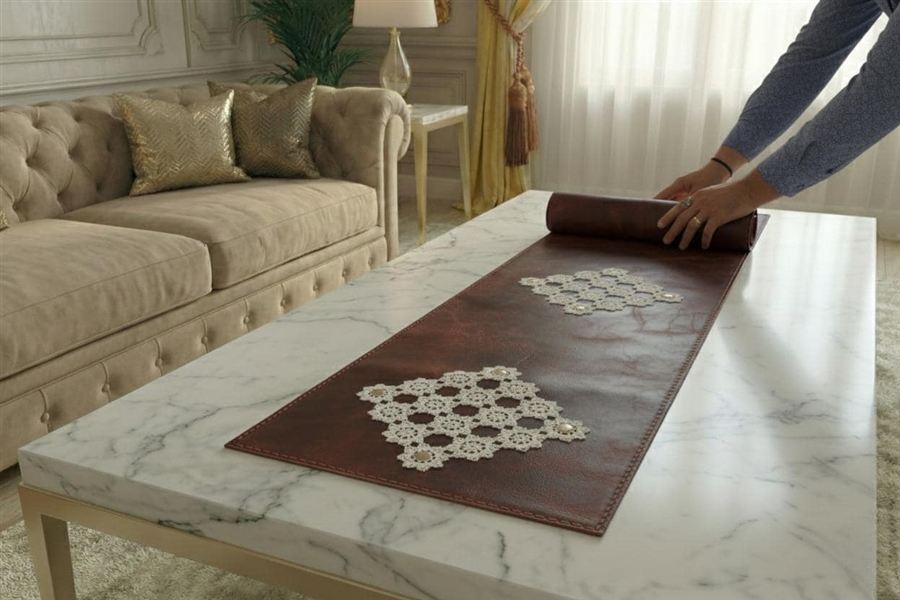 A hand-stitched oxblood full-grain leather table runner with two cream Turkish needle-lace motifs set near its ends, being unrolled on a white marble coffee table