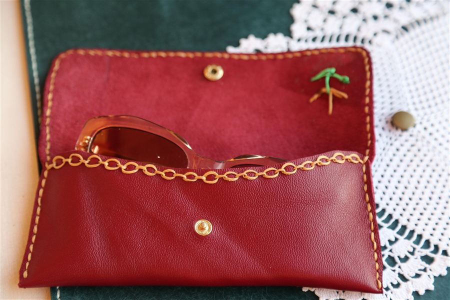 A hand-stitched leather eyewear case with embroidered detail, lying closed on a pale surface beside a pair of glasses