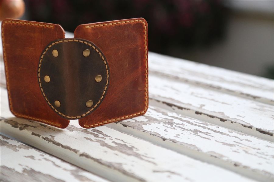 A hand-stitched leather wallet with an antique brass rivet set through the front panel, the surrounding leather smooth and undistorted, with cream saddle stitching along the edges