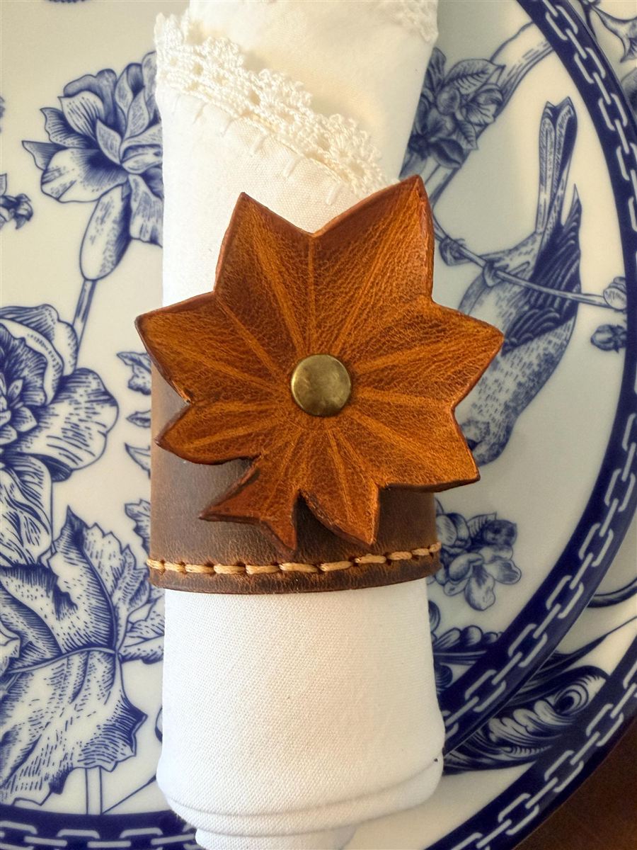 A hand-stitched leather napkin cuff with a maple-leaf motif and an aged brass rivet, holding a white lace-edged napkin on a blue-and-white patterned plate