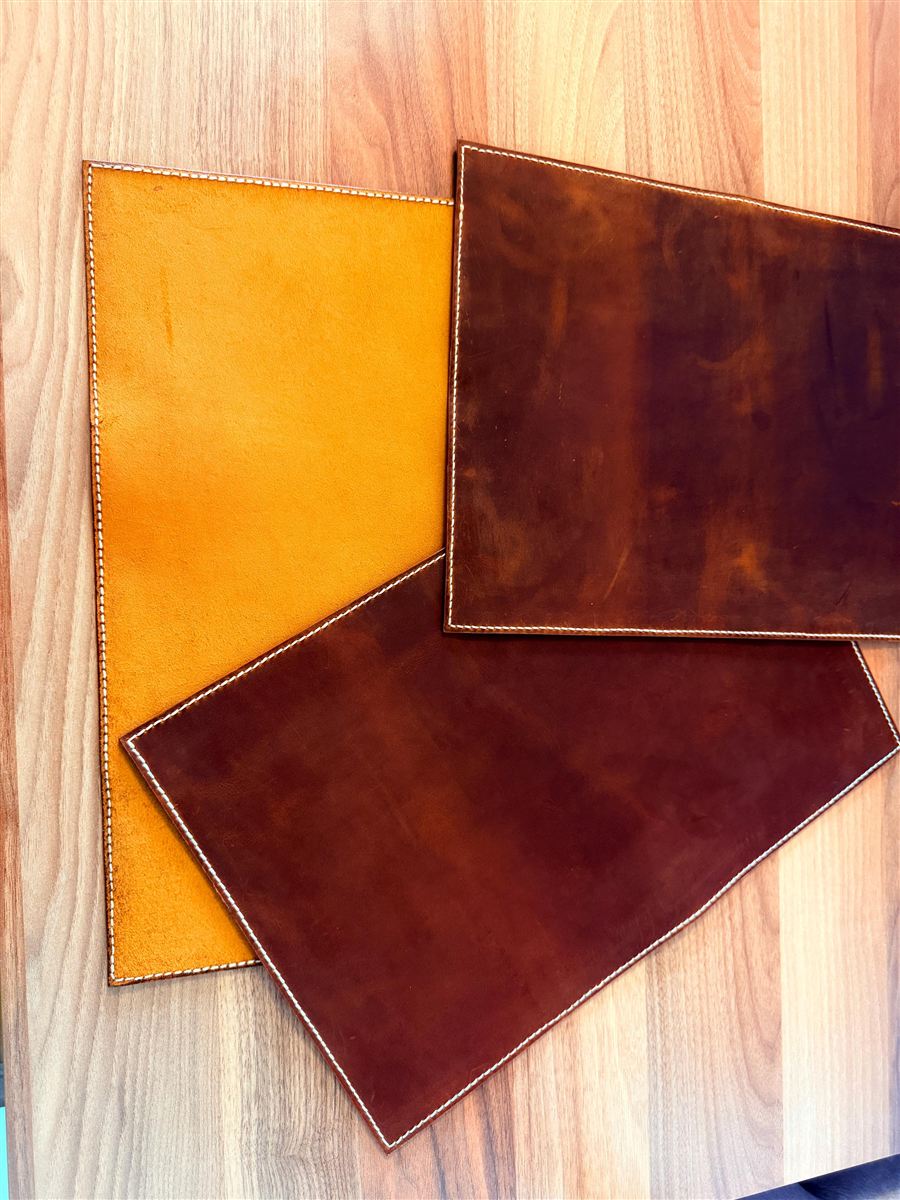 Three hand-stitched crazy horse leather placemats overlapping on a warm wooden table — one honey-tan, two deep cognac — each edge-stitched with cream thread