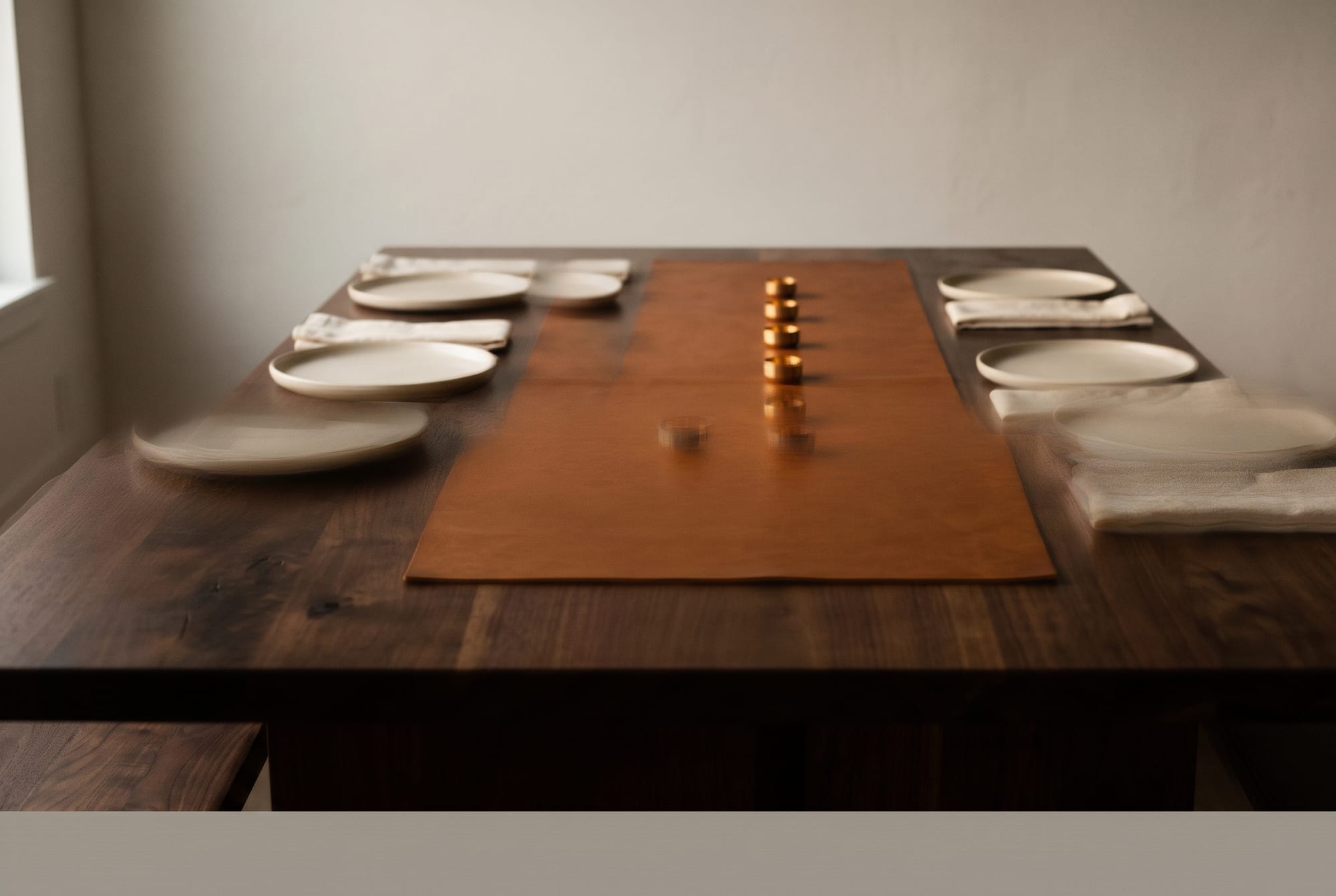 A cognac full-grain leather table runner laid down the centre of a dark walnut dining table, a third of its width and stopping short of both ends, with cream stoneware plates on the bare wood and a single line of small brass candle holders