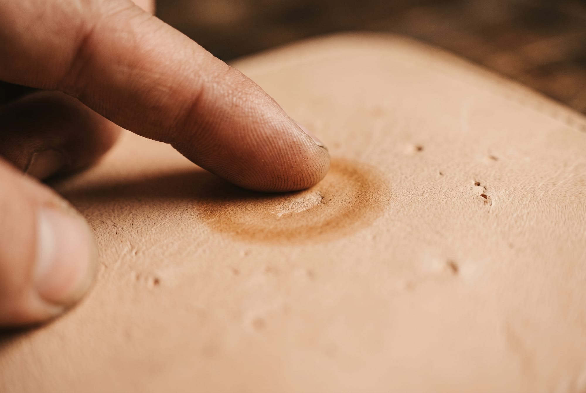 Close-up of a fingertip rubbing slow circles over a light scuff on natural veg-tan leather, half of the mark already blended back into the surface