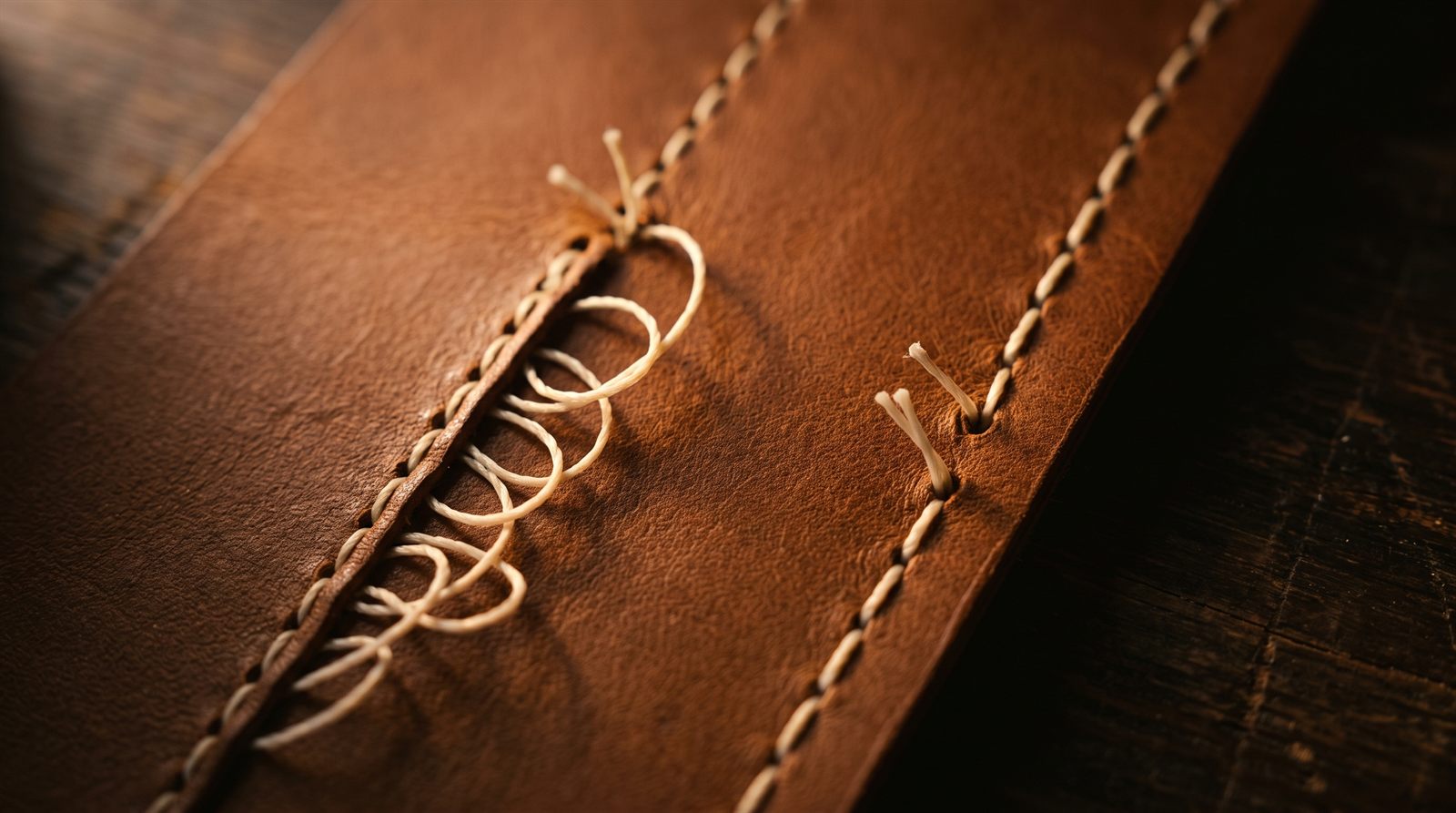 A machine-stitched leather seam laddering open in loose loops where one thread was cut, beside a saddle-stitched seam holding firm with a single missing stitch