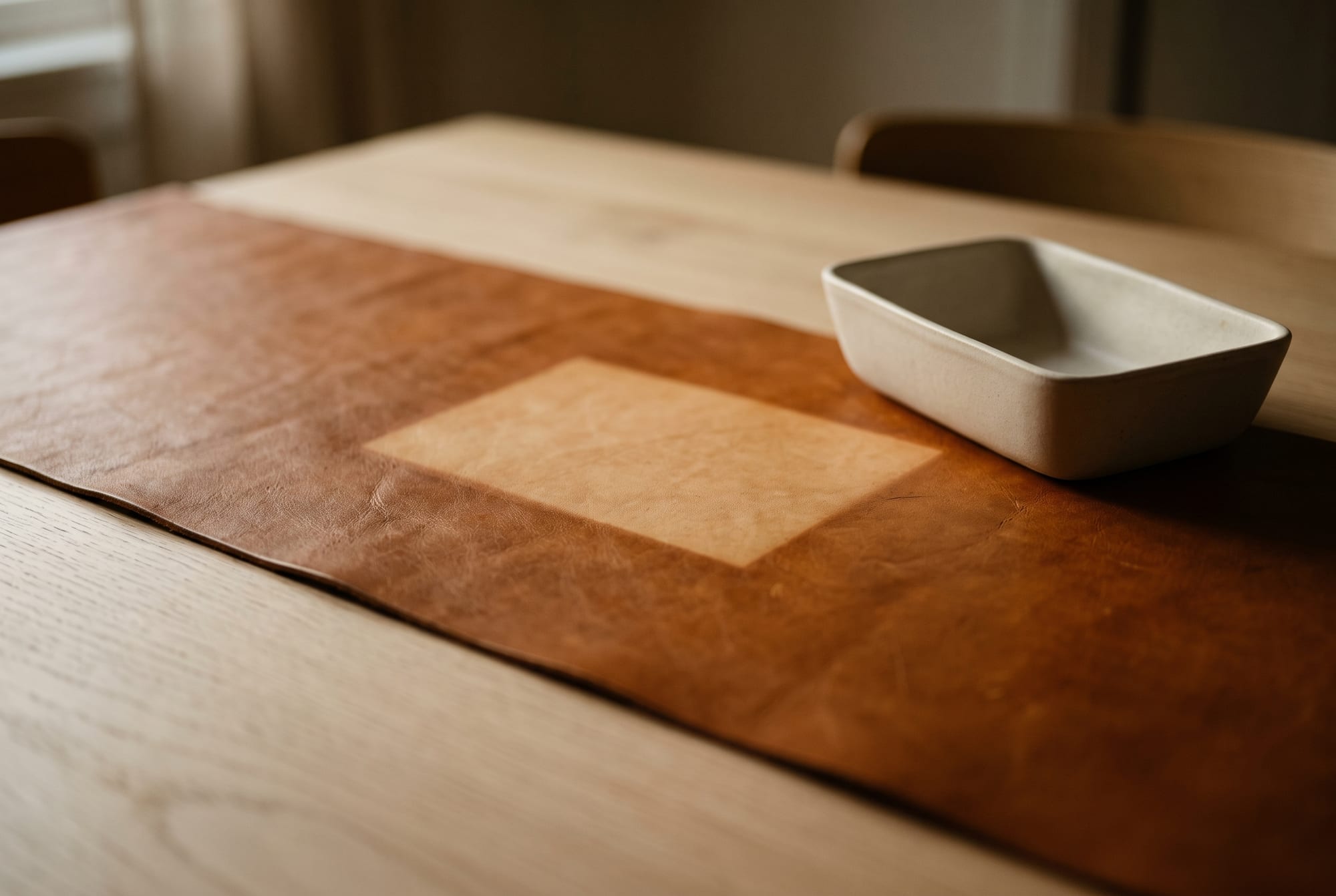 A cognac leather table runner on a dining table in low autumn window light, a hard-edged pale rectangle visible across it where a bowl has been sitting for months