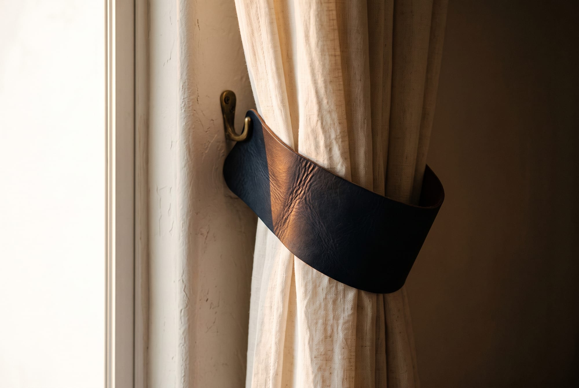 Navy blue leather curtain tiebacks holding a linen curtain beside a sunlit window, the knotted fold catching hard afternoon light while the inner face stays in shadow