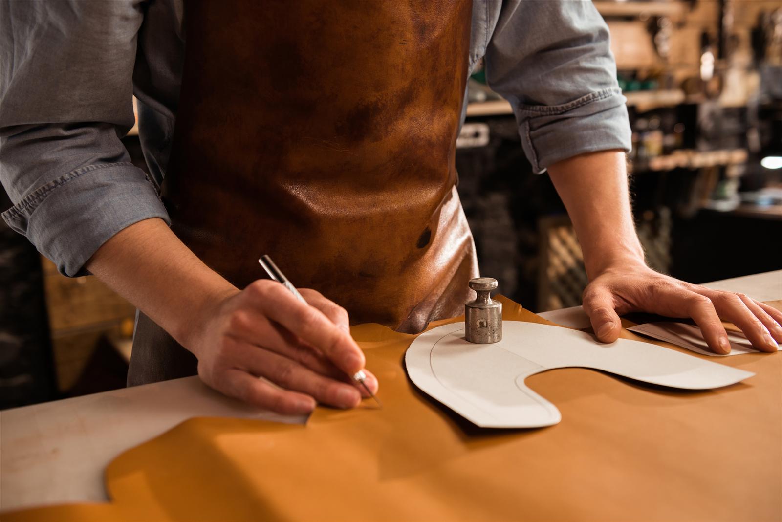 A pair of steel callipers closed on the edge of a cognac leather panel on a workbench, the jaws gripping the cut edge where the dense grain layer and the looser fibres beneath it are visible