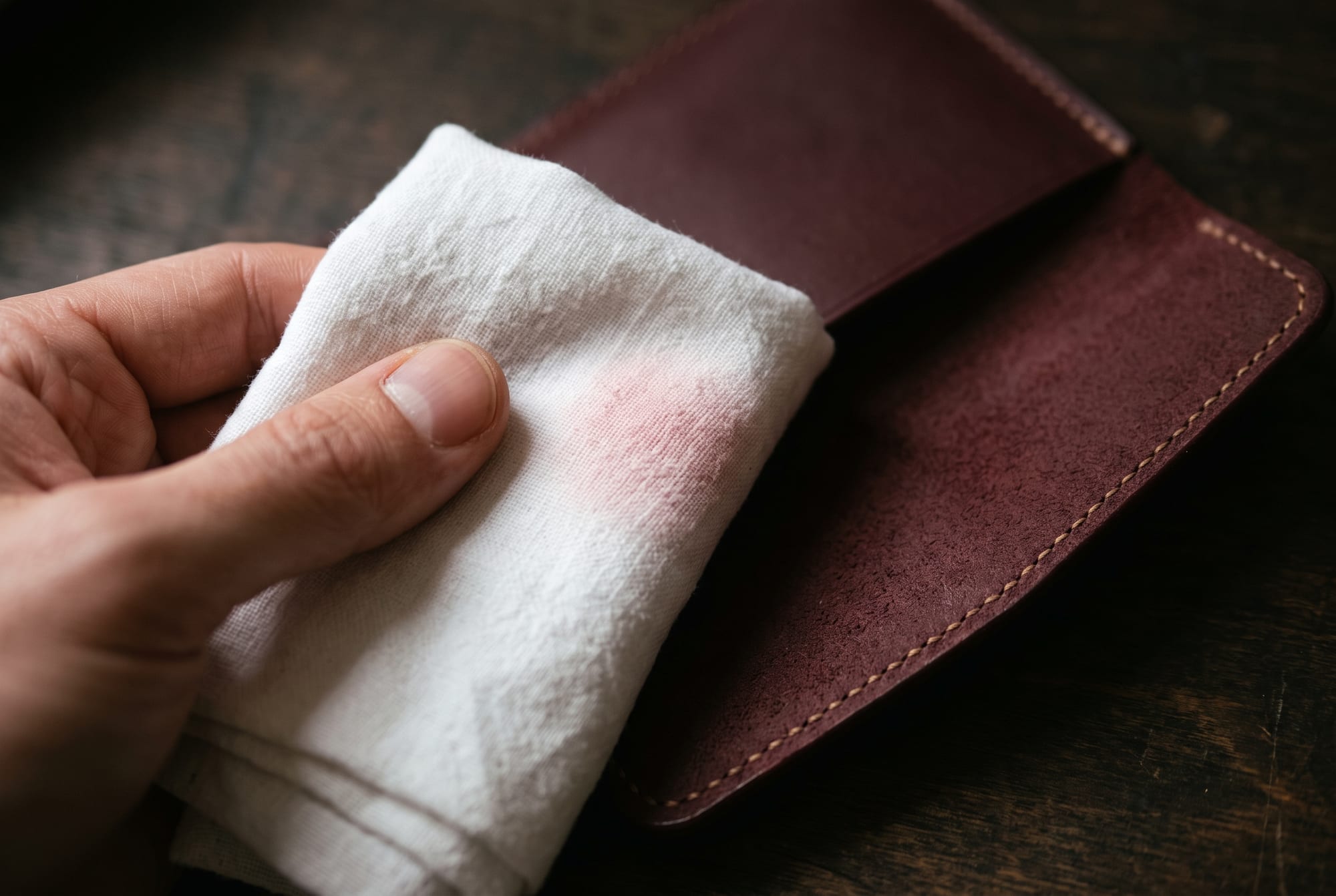 A folded white cotton cloth being pressed against the inside flap of a deep burgundy leather wallet, a faint pink blush visible on the cloth where it has made contact