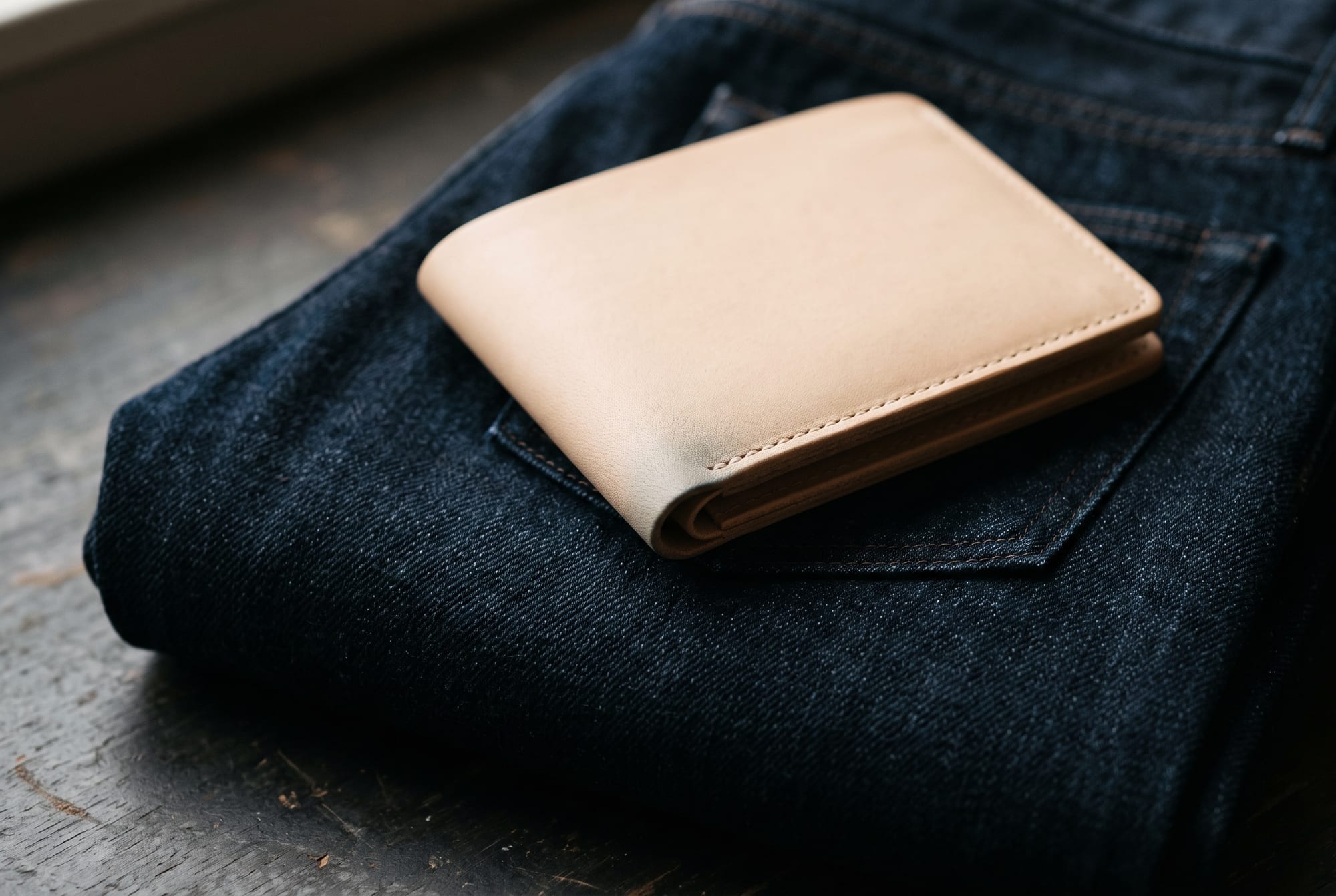 A pale natural vegetable-tanned leather wallet lying on folded dark indigo denim in low window light, a faint blue-grey shadow visible on the leather exactly where it has been resting