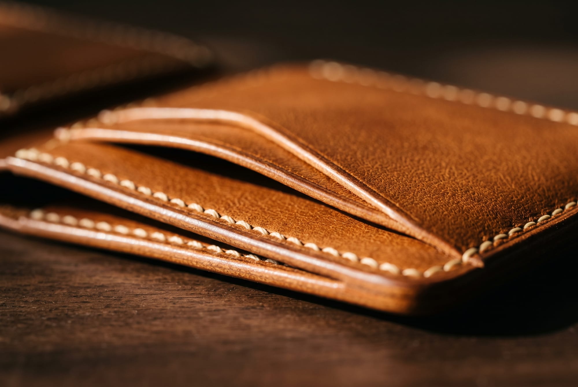 Raking light along the closed spine of a well-used cognac leather bifold, showing a broad soft curve darkened by years of handling with the saddle stitching running cleanly through the fold and no hard crease line