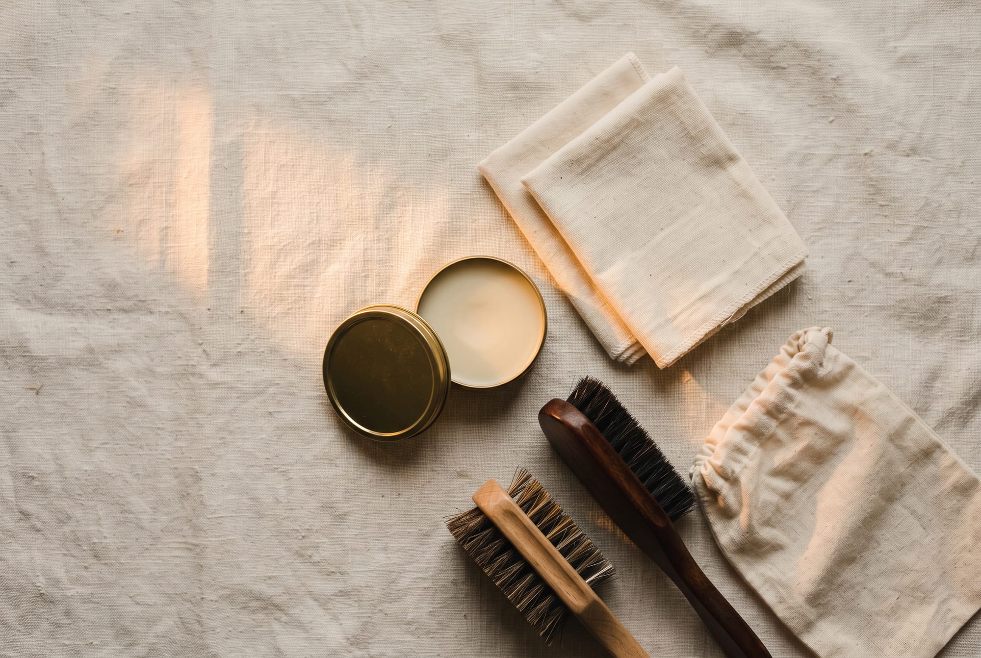 The honest leather care kit laid out on a workbench: a folded cream cloth, a small tin of beeswax conditioner, a horsehair brush and a dish of cornstarch