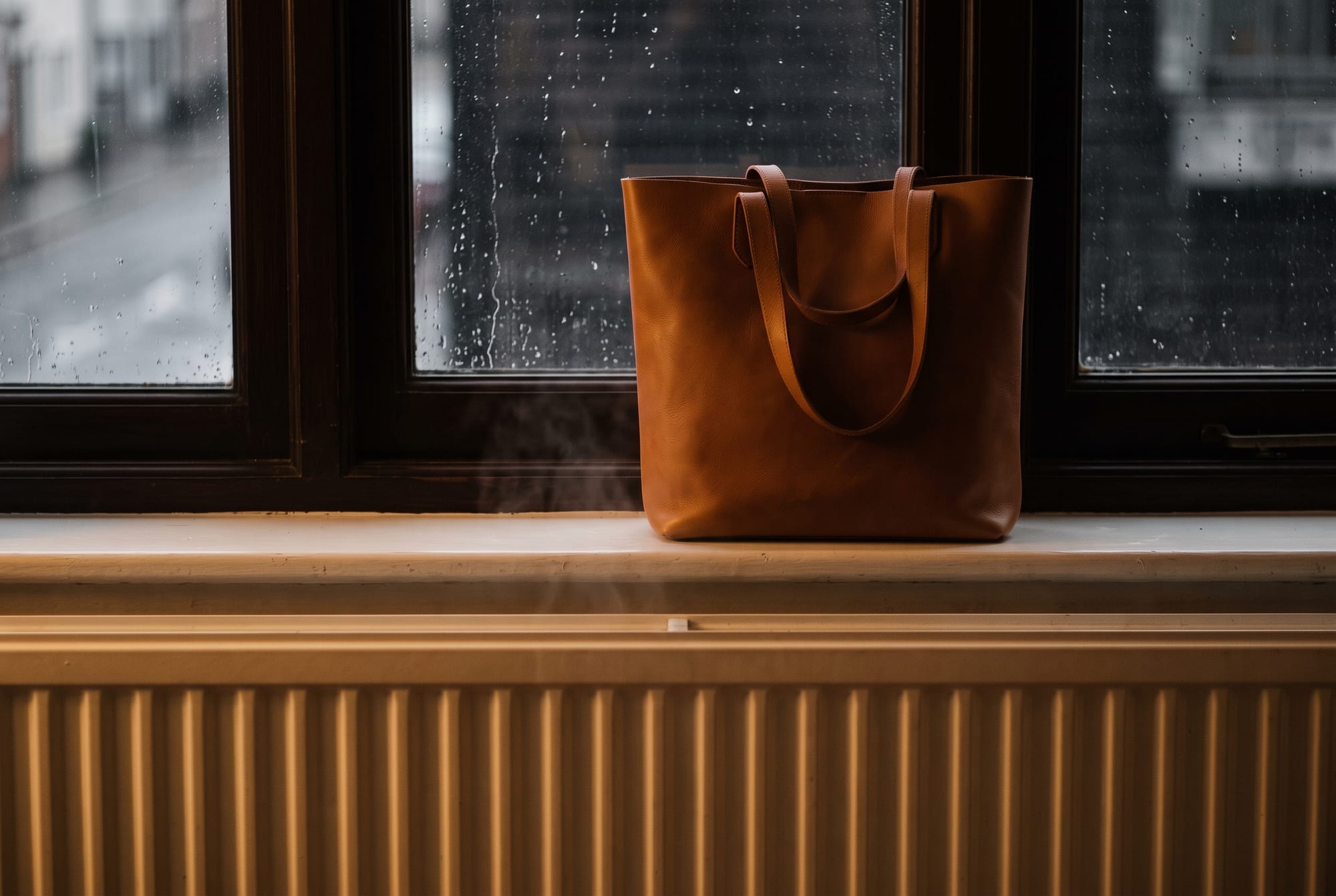 A cognac leather tote standing on a windowsill in low winter light, rain beaded on the dark glass behind it and the white ribs of a radiator just visible below the sill