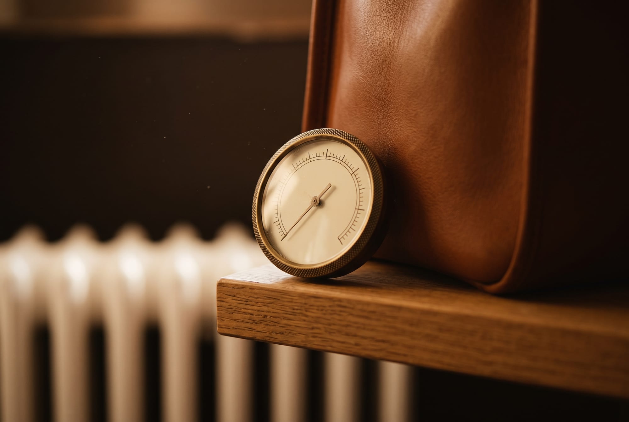 A small round hygrometer resting against a cognac leather bag on a wooden shelf, its needle low in the dry zone, with the ribs of a radiator out of focus below